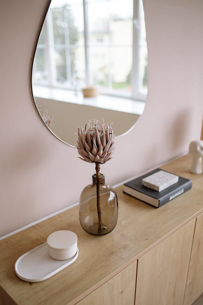 Modern interior with a wooden table, decorative mirror, and vase with dried protea flower.